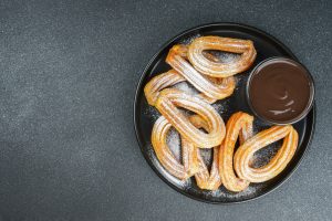 Traditional spanish dessert churros, fried dough pastry dusted with powdered sugar and chocolate sause on black background. Street food, sweet snack, homemade dessert. Top view, copy space